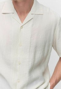 Chemise à manches courtes, couleur crème pâle, avec une apparence texturée, dotée d'un col notch et de boutons clairs sur le devant.
