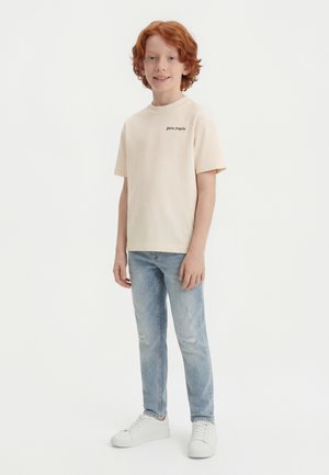 Garçon aux cheveux roux debout, portant un t-shirt beige Palm Angels, un jean bleu clair déchiré et des baskets blanches sur un fond blanc.