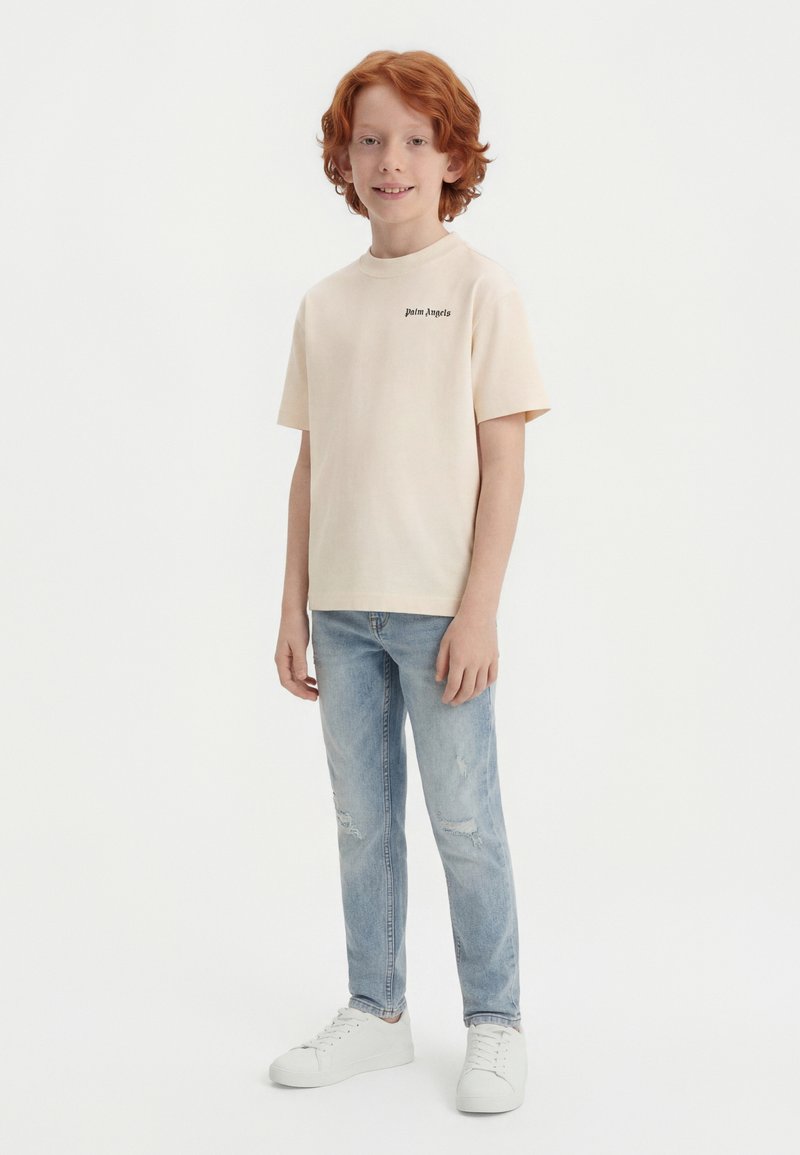 Κόκκινο αγόρι όρθιο, φοράει μπεζ μπλούζα Palm Angels, ανοιχτό μπλε σκισμένο τζιν και λευκά αθλητικά παπούτσια σε λευκό φόντο.