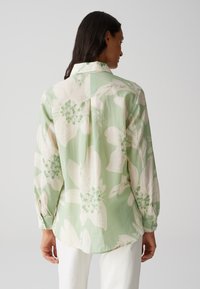 Camicetta floreale verde chiaro realizzata in tessuto morbido, caratterizzata da grandi fiori bianchi e accenti verdi, con colletto e maniche lunghe.