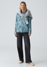 Camicia azzurra chiara con motivo floreale, abbinata a jeans a gamba larga in denim scuro e un maglione grigio a maglia posato sulle spalle. Sneakers casual.