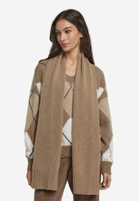 Beżowy, dziergany szalik o miękkiej fakturze, z prążkowanym brzegiem, prezentowany razem z wzorzystym swetrem w ciepłych odcieniach brązu i kremu.