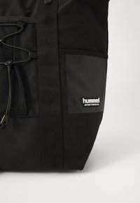 Sort sportstaske med netlomme og justerbar snor, med en "hummel SPORTSWEAR" logo-patch på forsiden.