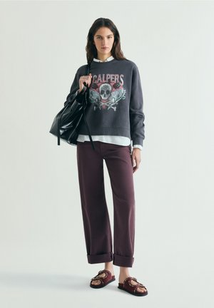 Frau steht und trägt ein dunkel bedrucktes Sweatshirt, ein weißes Hemd, hochgekrempelte weinrote Hose, Sandalen und hält eine große schwarze Handtasche über einer Schulter.