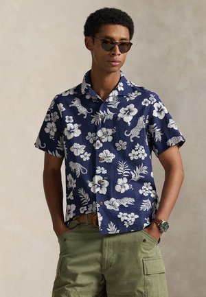 Chemise à manches courtes, bleu marine, à motifs floraux avec hibiscus blanc et imprimé léopard. Elle présente un devant à boutons et un col décontracté.