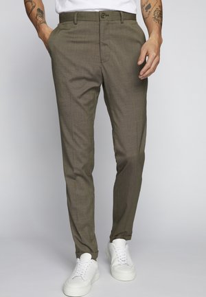 Pantalon slim ajusté vert olive porté avec des baskets blanches, mains détendues sur les côtés, tatouages visibles sur les avant-bras, fond uni clair.