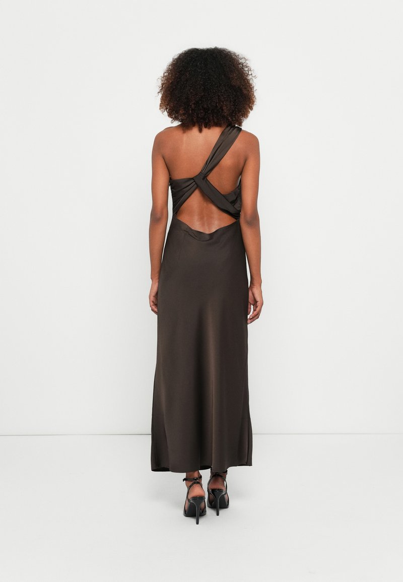 Robe en satin marron avec un design de bretelle torsadée, dos nu, coupe longueur sol et texture lisse et soyeuse. Portée avec des chaussures à talons noires.