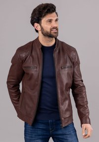 Brown leather jacket with a slim fit, zippered pockets, quilted shoulder detail, and a stand-up collar. Worn over a navy sweater.