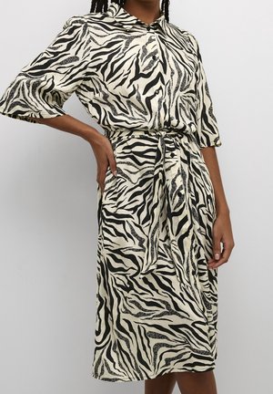 Kobieta w sukience do kolan z czarno-białym wzorem w paski zebry, krótkimi rękawami, guzikami i tkaninowym paskiem związanym w talii.