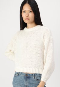 Cream-colored knitted sweater with a round neckline, loose fit, and ribbed hem. It features a textured, soft fabric and slightly puffed sleeves.