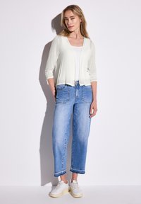 Lätt, långärmad cardigan i krämfärg över en vit linne, tillsammans med medelblå, löst sittande jeans och vita sneakers.