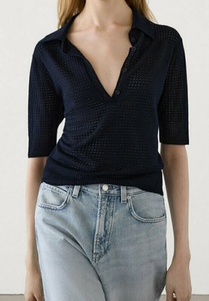 Woman wearing a black textured knit top with a deep V-neck and short sleeves paired with light blue high-waisted jeans.