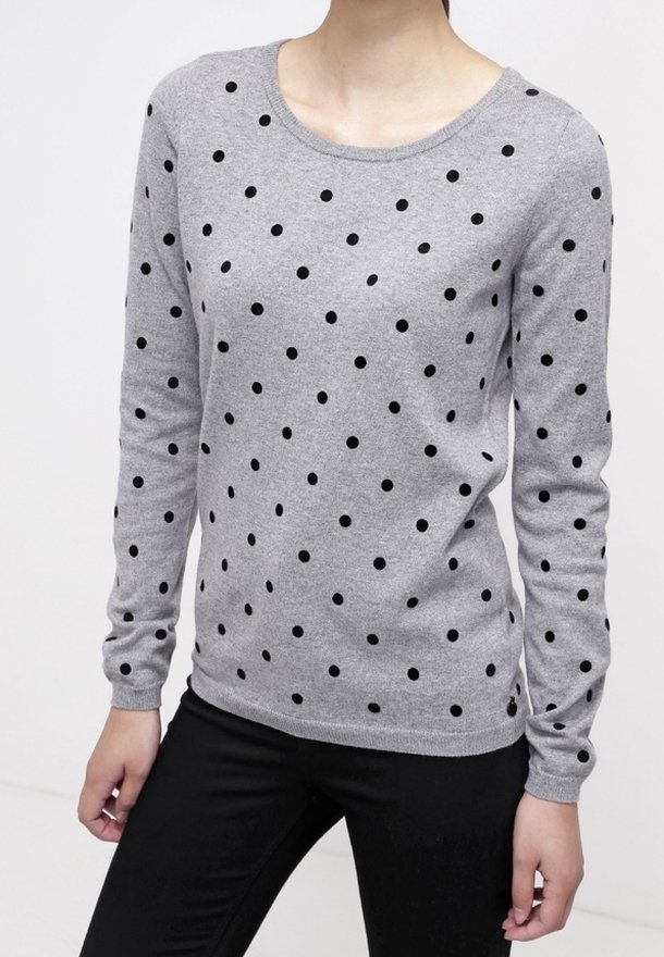 Femme portant un pull gris à manches longues avec des pois noirs et un pantalon noir, debout avec une main détendue et l'autre légèrement pliée.