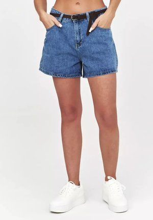 Kobieta w niebieskich szortach jeansowych z czarnym paskiem i białych trampkach na platformie, stojąca z rękami w kieszeniach na jednolitym tle.