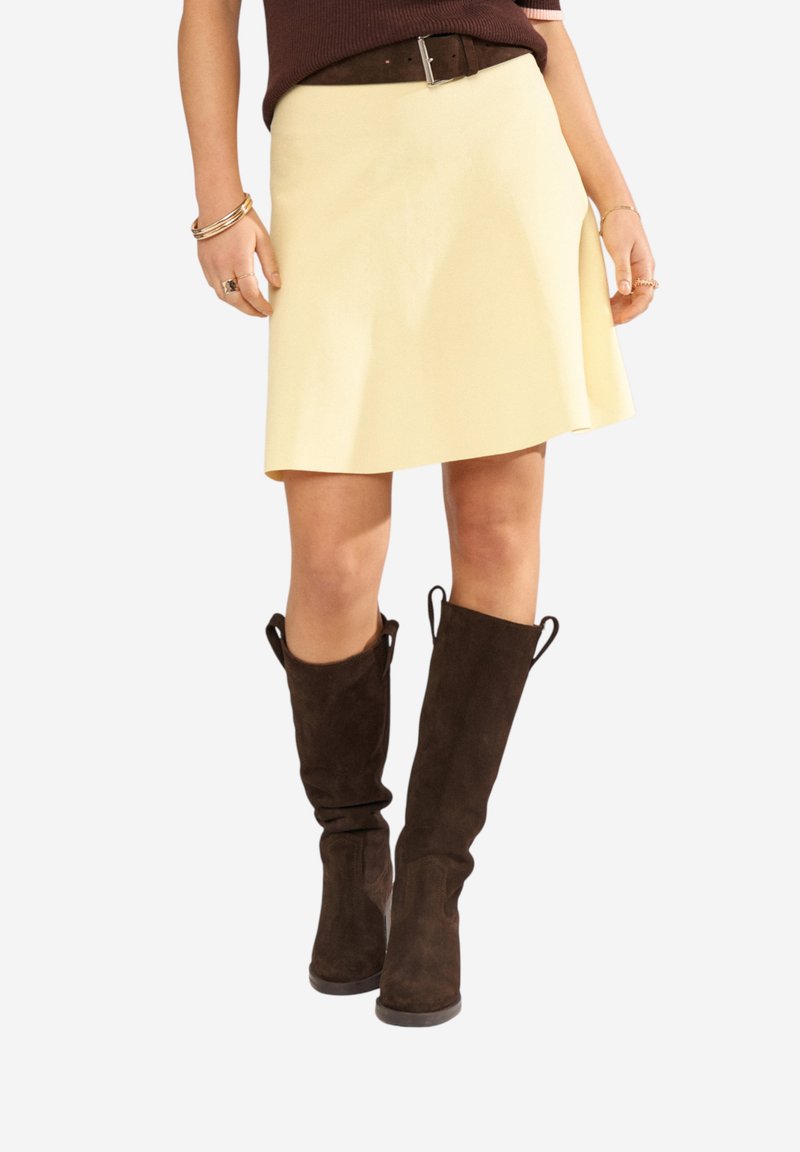 Femme portant une jupe trapèze crème, une ceinture marron, des bottes en daim marron foncé montantes jusqu'aux genoux et des bracelets dorés aux deux poignets.