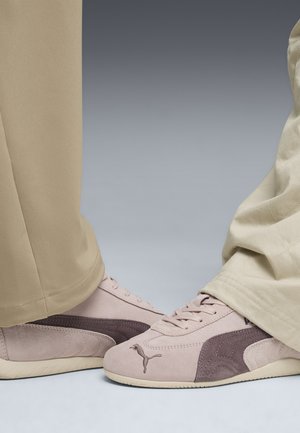 Zapatillas Puma en rosa claro y ante marrón con diseño de cordones. Acentos texturizados y logotipo visible. Unos pantalones de tela beige enmarcan los zapatos.