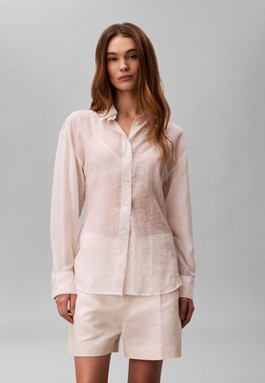 Młoda kobieta z długimi brązowymi włosami, ubrana w przejrzystą białą koszulę na guziki i jasnobeżowe szorty na jednolitym szarym tle.