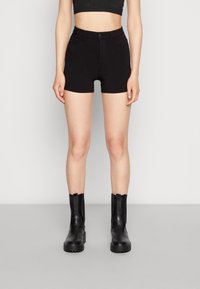 Pantalones cortos de mezclilla negros con cintura alta, diseño ajustado y sin bolsillos. Llevados con botines negros que tienen una suela gruesa y una costura distintiva.