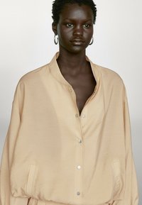 Ljusbeige oversized skjorta i mjukt tyg, med ståkrage och silverfärgade tryckknappar, sidofickor och avslappnade ärmar.