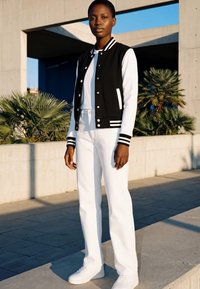 Ženska, oblečena v črno-belo varsity jakno, bele hlače in bele superge, stoji na prostem blizu betonske strukture in zelenja.