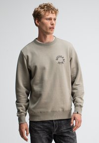 Grijze katoenen sweatshirt met ronde halslijn en ribgebreide manchetten. Voorzien van een vooraanstaand logo in donkergrijs. Gepaard met een donkere spijkerbroek.