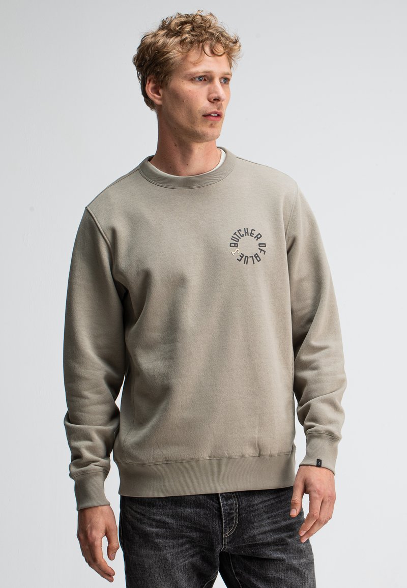 Grijze katoenen sweatshirt met ronde halslijn en ribgebreide manchetten. Voorzien van een vooraanstaand logo in donkergrijs. Gepaard met een donkere spijkerbroek.