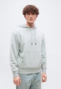 Νέος άνδρας με σγουρά μαλλιά, που φοράει ανοιχτό γκρι φούτερ με κουκούλα με το κείμενο "BOSS" και ανοιχτό μπλε τζιν, στέκεται μπροστά σε απλό λευκό φόντο.