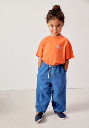 Niño pequeño con cabello rizado recogido en un moño, vestido con una camiseta naranja y pantalones azules sueltos, de pie con las manos en los bolsillos sobre un fondo liso.