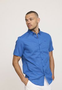 Chemise à manches courtes, boutonnée, en tissu bleu vif. Elle présente un col, des poches avant et une finition légèrement texturée. Les boutons sont de couleur bleu foncé.