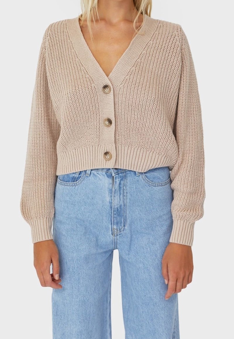 Béžový pletený cardigan s hlubokým výstřihem do V, zdobený třemi velkými knoflíky, žebrovanými manžetami a zkráceným střihem, doplněný světle modrými džíny s vysokým pasem.