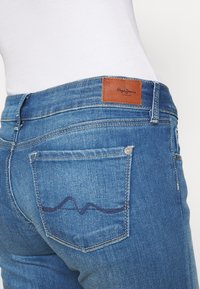 Vaqueros de mezclilla en un lavado azul medio con un parche de cuero con el logo de Pepe Jeans en la parte trasera. Presenta un bolsillo trasero con un detalle de costura en patrón de ola.