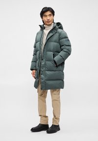 Vihreä puhallettava takki huputtuna, tikattu pinta, etupuolen nepparipainike, sivutaskut, yhdistettynä beigen väriseen villatakin ja vaaleanruskeisiin housuihin.