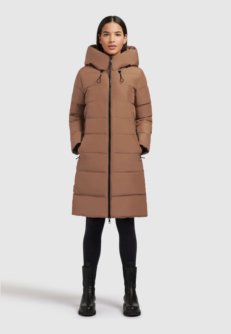 Brun pufferjakke med hætte, der har en fuld frontlynlås, sidelommer og vandret syninger for ekstra struktur. Sorte støvler fuldender looket.