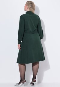 Abito verde scuro con maniche lunghe, colletto alto e gonna svasata. Presenta una vita evidenziata da una cintura e scarpe con tacco argentate. Tessuto morbido.