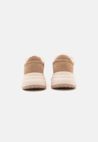 Ζευγάρι μπεζ slip-on παπούτσια με χοντρές λευκές σόλες και μαλακή, τριχωτή επένδυση, εμφανίζονται από πίσω σε λευκό φόντο.