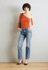 Top arancione asimmetrico con manica lunga da un lato, abbinato a jeans azzurri chiari e décolleté nere a punta, su un pavimento in legno.