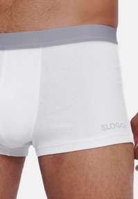 Weiße Baumwoll-Boxershorts mit einem grauen elastischen Bund, der das "SLOGGI"-Logo in Grau trägt. Der Stoff hat eine glatte Textur.