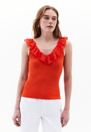 Top sin mangas de un brillante color naranja, con escote en V profundo y borde de volantes rojos texturizados a lo largo del cuello. Tela suave y ajustada. Pantalones blancos.