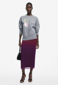 Pull en maille gris avec des détails en sequins, associé à une longue jupe violette transparente. Le mannequin porte des talons noirs et tient un petit sac à main noir.