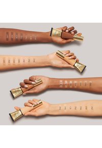 Fondsbuisjes vastgehouden door armen die verschillende huidtinten tonen. Elke arm toont aangebrachte foundationkleuren met genummerde labels ter referentie.