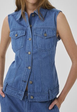 Kvinde iført ærmeløs blå denimvest med metalknapper og forlommer med klap, kombineret med matchende denimbukser.