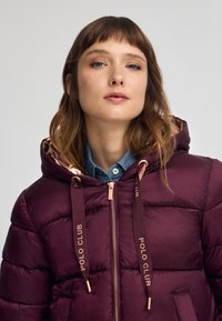 Burgundy pufferjacka med huva, gul dragkedja och snoddar med logotryck. Har sydda sektioner för isolering och sidofickor.