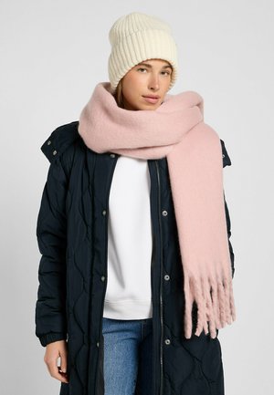 Sciarpa di lana rosa chiaro avvolta attorno al collo, con estremità frangiate, abbinata a un maglione bianco e a un berretto a coste crema. Giacca nera trapuntata.