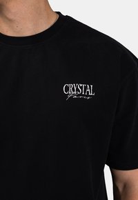 Zwarte katoenen t-shirt met witte geborduurde tekst "CRYSTAL Paris." Korte mouwen en een ronde hals. Gladde textuur.