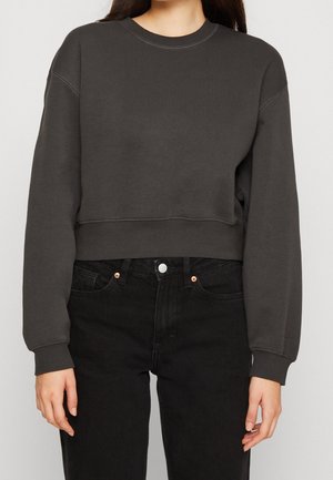 Frau trägt einen dunkelgrauen Crop-Sweatshirt und hoch taillierte schwarze Jeans, steht mit entspannten Armen an den Seiten.