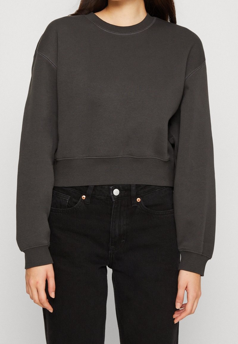 Femme portant un sweat-shirt court gris foncé et un jean noir taille haute, debout, les bras détendus le long du corps.