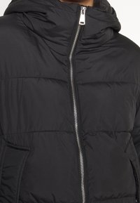 Czarna puchowa kurtka z kapturem, posiadająca srebrny zamek błyskawiczny i pikowany wzór, z bocznymi kieszeniami i dopasowanym krojem.