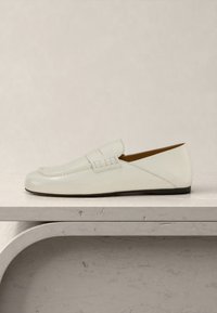 Λευκό δερμάτινο μοκασίνι χωρίς κορδόνια με χαμηλή μαύρη σόλα, μύτη μοκασίνι και λεπτομέρεια με λουράκι πάνω από το πάνω μέρος, τοποθετημένο σε φωτεινή πέτρινη επιφάνεια.