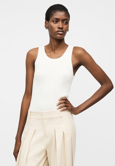 Γυναίκα που φοράει λευκό αμάνικο κοντόπλεκτο τοπ με ριπ υφή και μπεζ πλισέ παντελόνες με ψηλή μέση, στέκεται με ένα χέρι στη μέση.