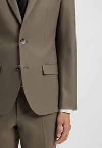 Brown tailored suit jacket with two buttons, lapels, and a breast pocket. Smooth fabric with a slight sheen; hand resting on the jacket.